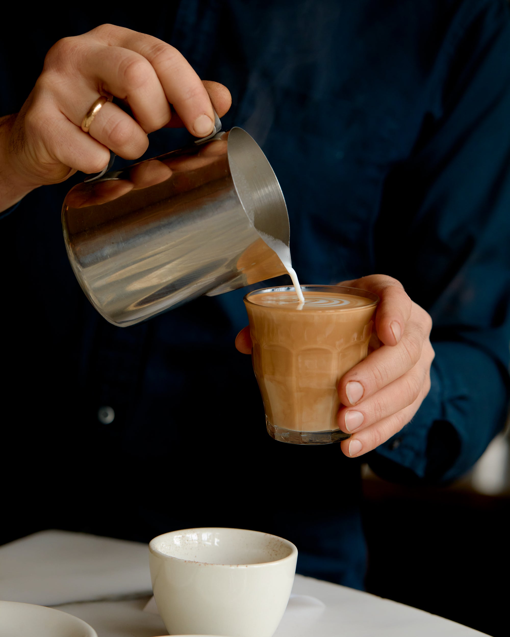 A close-up of a person pouring steamed milk from a stainless steel pitcher into a glass of iced coffee or latte, with a white cup on a table in the foreground.